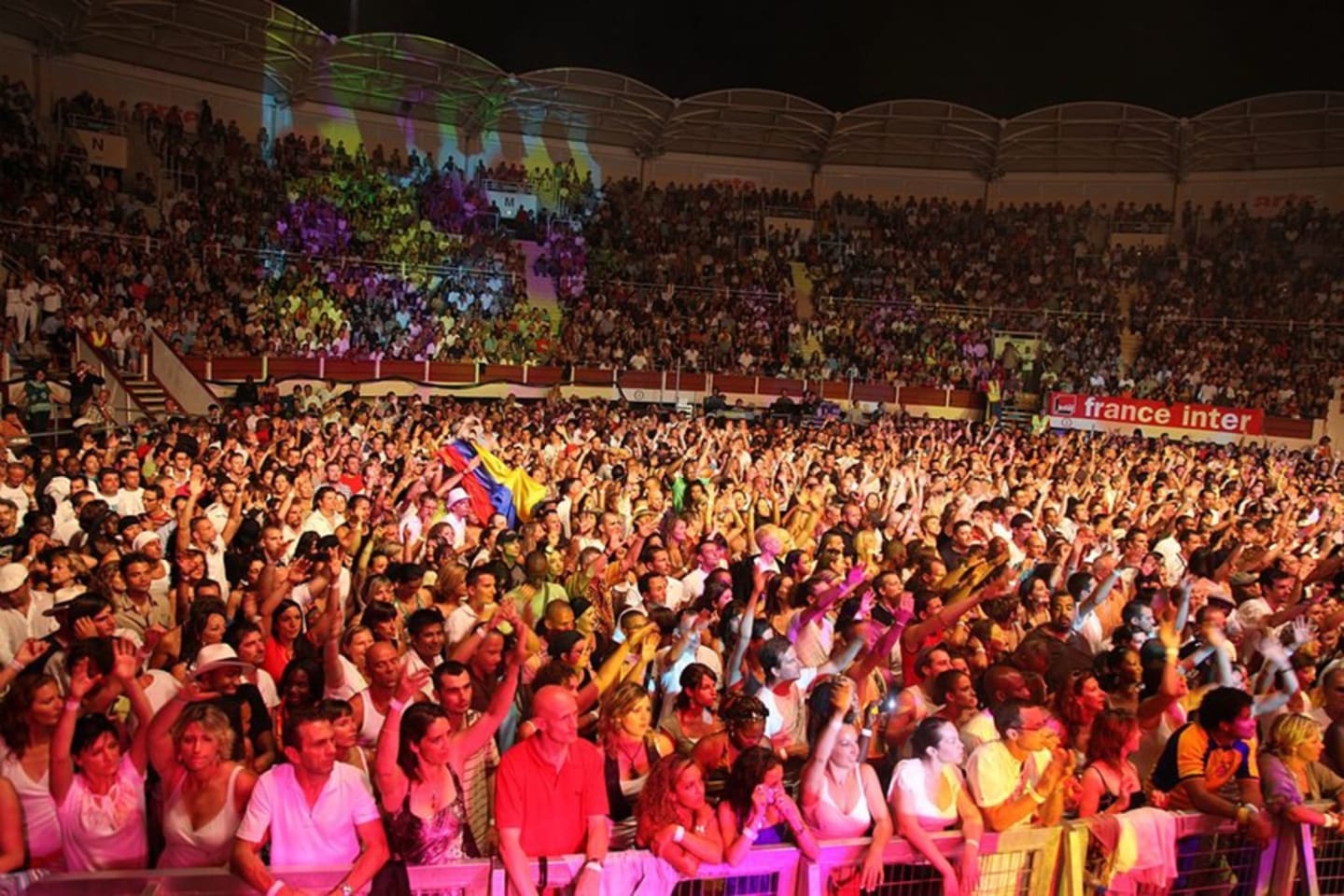 Foule impressionnante dans l'arène de corrida de Montréjeau, illuminée de couleurs vives et décorée de drapeaux, ambiance festive nocturne