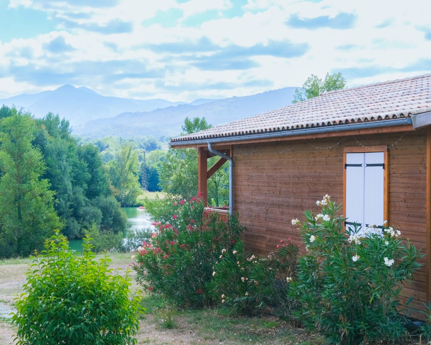 Chalet en bois aux portes blanches surplombant le Lac de Montréjeau à Clarac, entouré de plantes fleuries et de montagnes