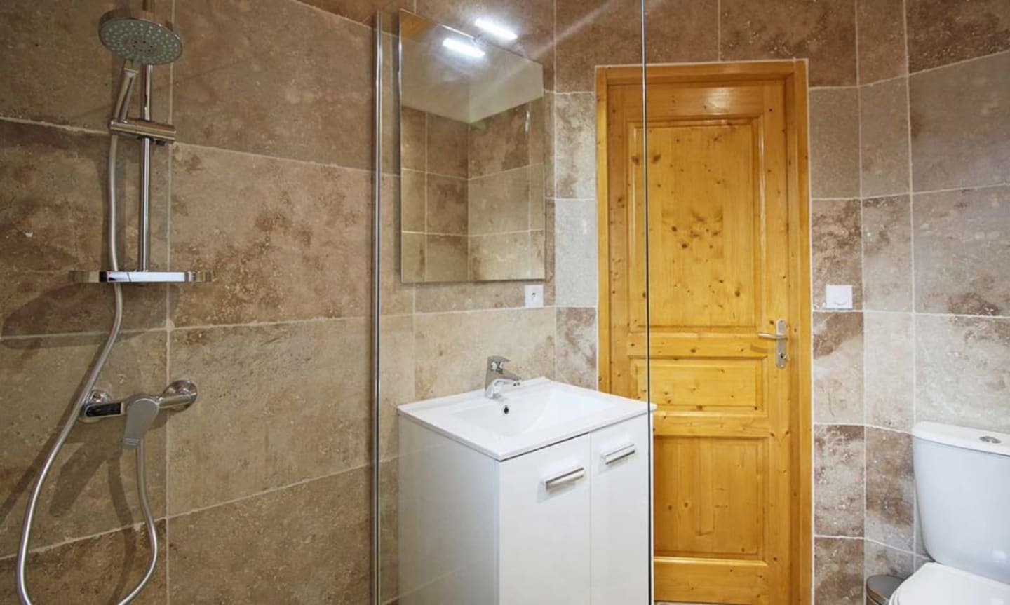 Salle de bain moderne aux Chalets Du Lac De Montréjeau avec douche à l'italienne, vasque blanche, porte en bois et travertin beige