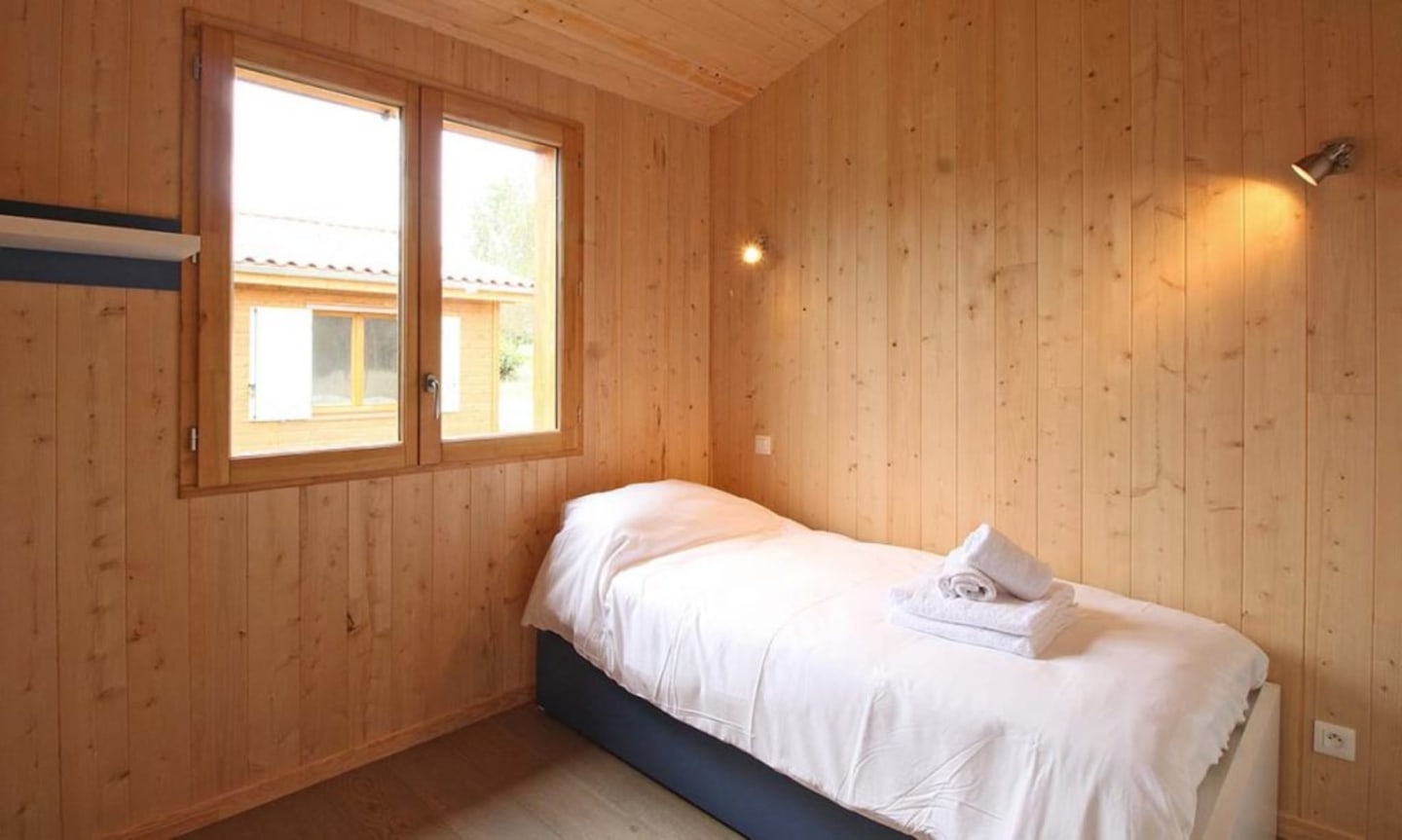Chambre douillette du chalet en bois avec lit simple, literie blanche et grandes fenêtres aux Chalets Du Lac De Montréjeau, Clarac