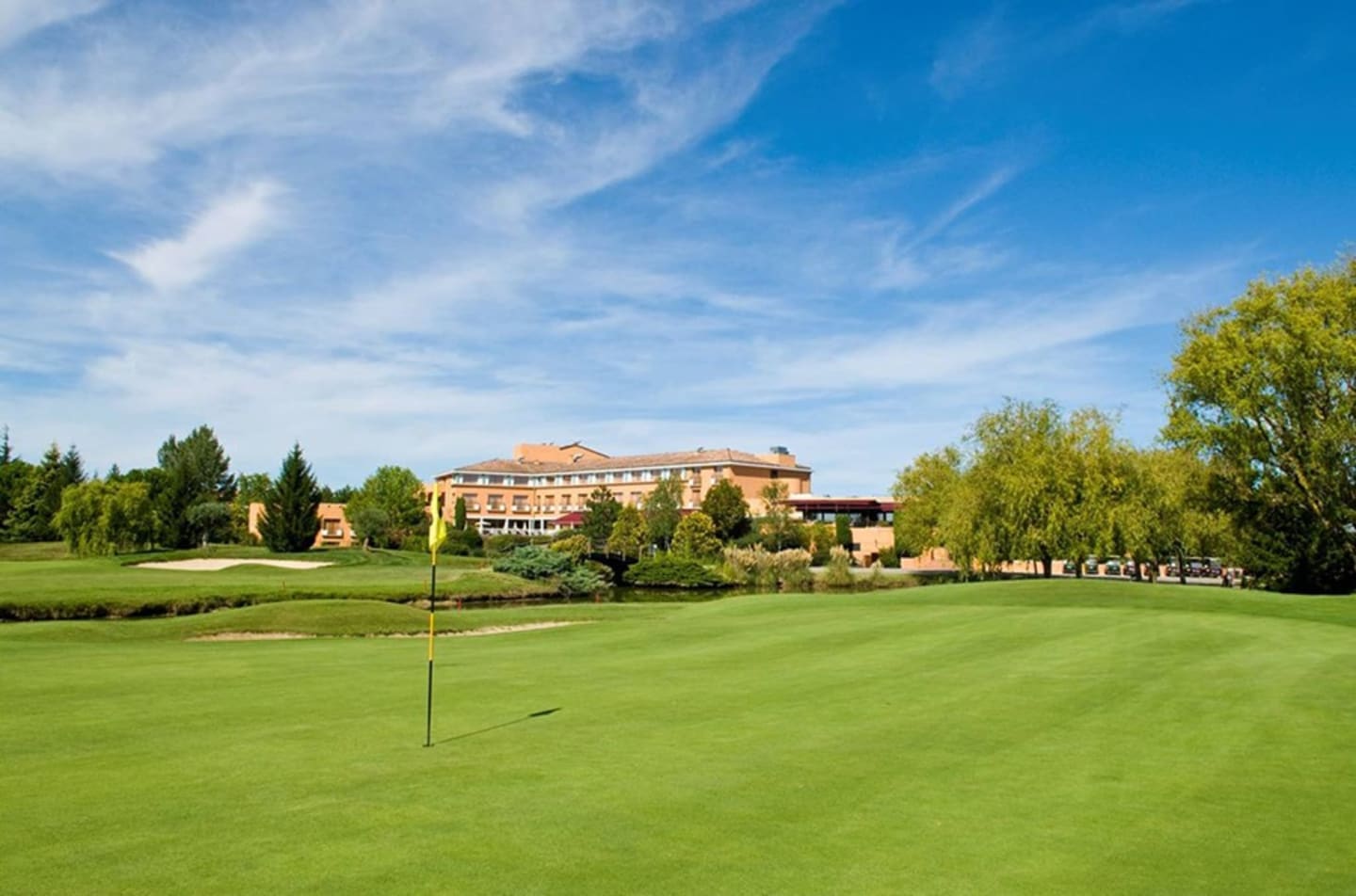 Golf course green with terracotta hotel building, trees and blue sky at Les Chalets Du Lac De Montréjeau, Clarac